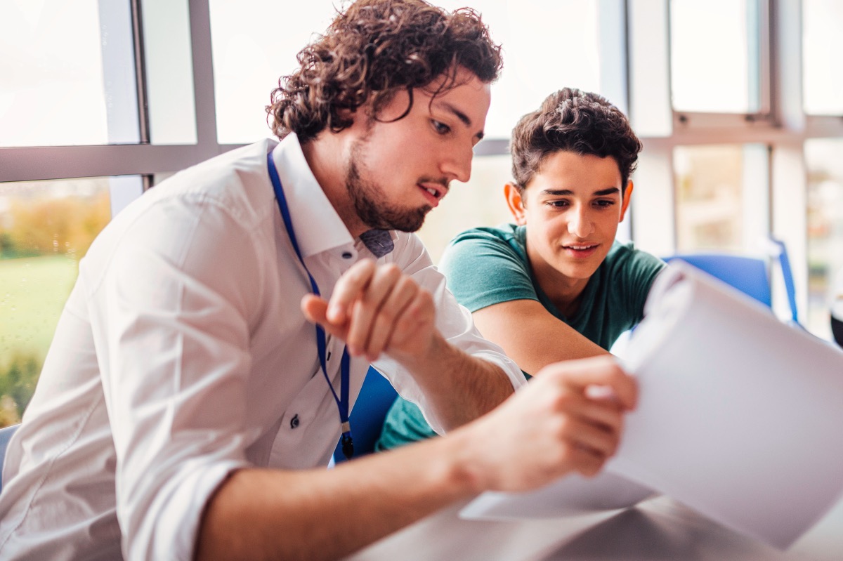 Teacher showing a student some results on his paperwork as they sit side by side at a table.