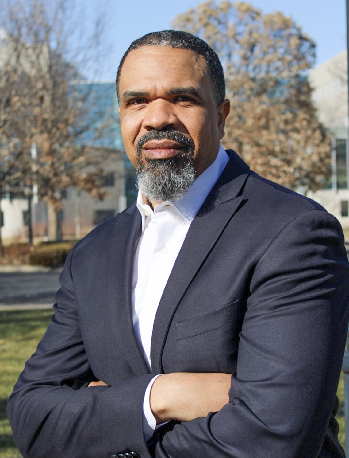 Headshot image of Dr. Allen standing on college campus wearing a suit looking into the camera